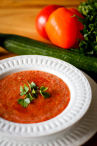 A white bowl on a white plate filled with tomato soup, granished with parsley, and a cucumber and tomatoes in the background