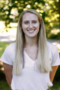 A smiling, white, long haired blonde female in a white shirt standing outside 