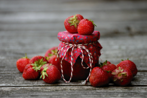 Strawberries laying around a glass jar of strawberry jam with a red fabric lid tied with twine.